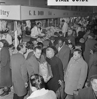 Black and white photograph of a busy indoor market at the Bull Ring shopping centre. Shoppers wearing coats are crowded around a market stall which is selling poultry products.