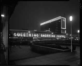 Black and white photograph showing part of the Bull Ring lit up at night, with a bright sign that reads ‘BULL RING SHOPPING CENTRE’. The office block rises in the background.
