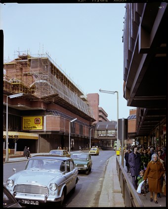 Colour photograph of a street view with active traffic and pedestrians holding shopping bags. Wood Green Shopping City is partially under construction, with scaffolding around the housing complex above the shop units.