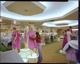 Colour photograph showing the interior of the D H Evans store at Wood Green Shopping City. There are large lights on the ceiling, a shop worker adjusting a mannequin dressed in pink, and multiple clothing racks in the background.