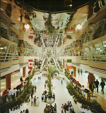 Colour photograph showing the interior of Eastgate Shopping Centre. There is a large reflective ceiling and shoppers, plants and escalators in the mall.