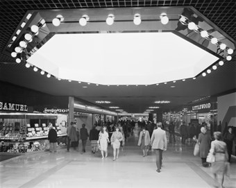 Black and white photograph showing the interior of Queensgate Shopping Centre. Shoppers are walking past the shop units, including Littlewood and H Samuel. Above is a huge octagon-shaped roof light.