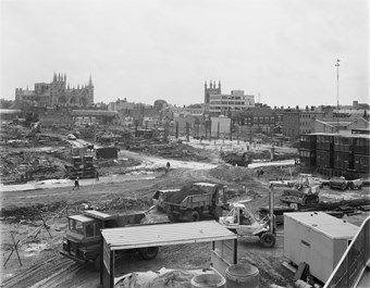Black and white photograph showing the site for Queensgate shopping centre at an early stage of construction. There are various construction vehicles and builders at work, and Peterborough Cathedral is in the distance.