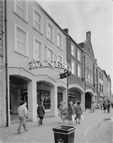 Black and white photograph showing an entrance to The Lanes Shopping Centre with a sign above. Pedestrians are walking along the street.