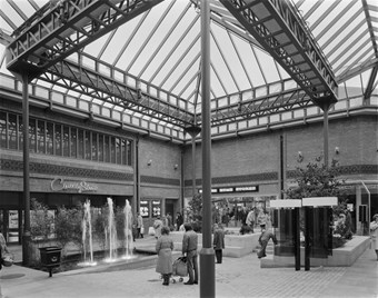 Black and white photograph showing shoppers in Central Square at The Lanes Shopping Centre. There are fountains, plants, and two telephone booths. The County Store and British Home Stores are in the background.