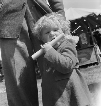A toddler with curly blond hair holds a stick of rock to her mouth. The child wears a long-sleeved coat and stands in front of the legs of a man. The image is in black and white.