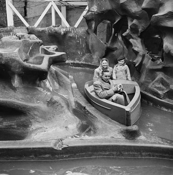 A boat travels along the channel of a water ride. In the boat, a smiling woman sits beside a young boy. A man sits in the prow and wipes his hands with a handkerchief. To the left is a false rocky outcrop of the ride. The image is in black and white.