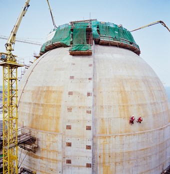 The image depicts a large dome which is white with specks of orange colouring. At the top, there is green netting and scaffolding. To the left there is a large yellow crane. On the dome itself, there are two people who are abseiling.