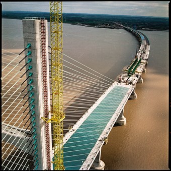 A view of the top of a bridge, which spans out to the coastline in the distance. The bridge is not yet finished. The river underneath it is a muddy-brown colour.