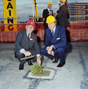 Two men wear suits and crouch down to plant a small tree. They are both wearing hard-hats. In the background, there are three other men. To the left of the picture, there is a large vertical sign saying ‘Laing’.