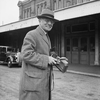 An old man smiles to the camera. He is holding a camera and wears and overcoat and hat. In the background there is a building and an old car. The image is in black and white.