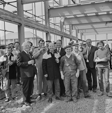 A crowd of men pose for a picture. Some are wearing work-overalls and donkey jackets and a few of the men wear suits. Some of the men are holding pints of beer. They look to be celebrating inside a large building which has scaffolding on the outside of it.
