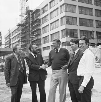 Five men pose for a photograph; all wearing smart clothes. The man in the middle shakes the hand of the man to his left. They are all smiling. In the background, there is a building site.