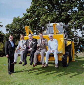 Five men in suits pose for a photograph. One man stands to the left of the picture. The other four sit in the digger of a JCB. In the background there's a tree.