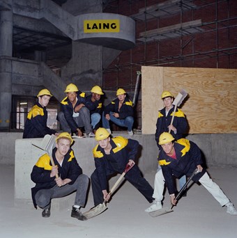 Eight men wear black and yellow donkey jackets and pose for a photograph. Each man holds a shovel as if they were doing a dance move. They pose in a dark room and it looks like it is underground. In the background, the Laing logo can be seen.