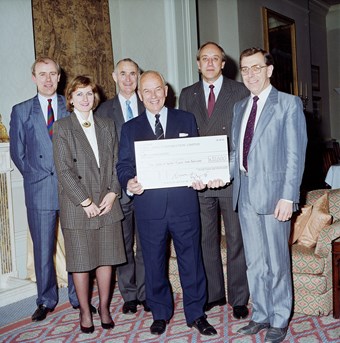 Five people pose for a photograph. There are four men and one woman. The man in the middle holds a large cheque. The photograph is in colour and all the people are smartly dressed.