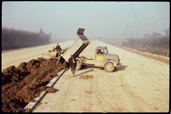 A long road recedes into the distance. A truck dumps a load of earth in the middle of the road, where a worker drives a small digger. The truck has a ‘Laing’ sign on it. Another worker stands next to the truck. He has an L on his back.