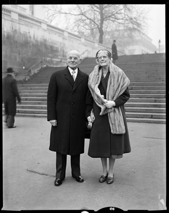 A smartly dressed man and woman pose for this black and white photo. The man wears a suit and the woman wears what looks to be a fur scarf. In the background there's a wide set of stone steps and a leafless tree.