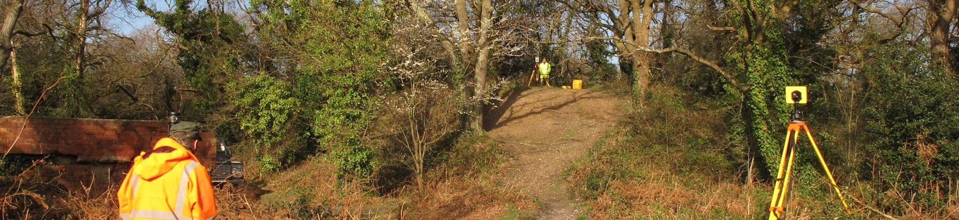 Two archaeologists in hi-vis gear surveying a small motte and bailey castle.