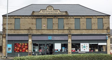Shop front with windows blocked up on first floor and the words 'Erected 1886 Drighlington Co-operative Industrial Society' carved in stone plinth above the gutter