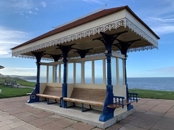 Shelter with ornate rooftop, blue pillars, and a wooden bench. Sea is visible in the background