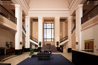Grand foyer with first floor walkway supported by columns, descending to wide central staircase in front of a large window and flanked by columns.