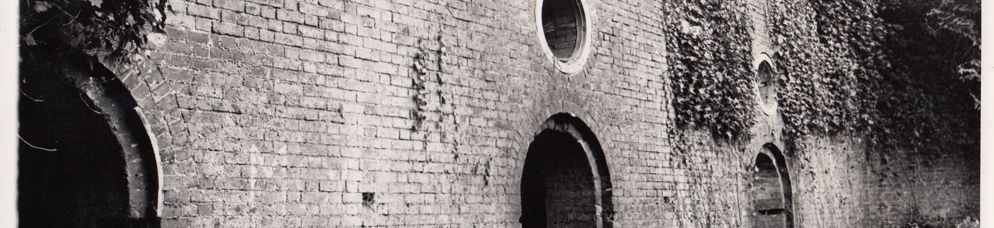 Black and white photo of high brick casemates with arched openings at ground level and circular windows.