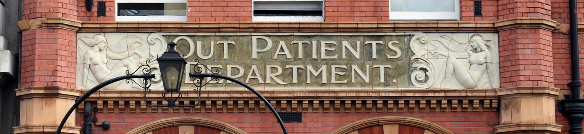 Entrance with the words 'Out Patients Department' engraved in the stone flanked by two women on either side. A lantern above the entrance gate partly obscures the view of the stone carving on the left.