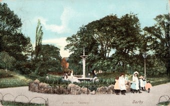 A group of women and children are gathered on a track in the right foreground, next to a fountain centre. There are trees in the background.