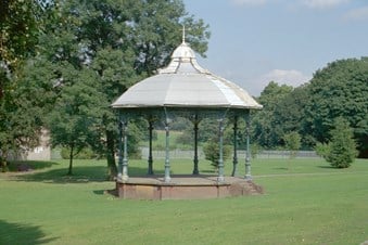 A bandstand in the foreground, surrounded by lawns with trees in the background.