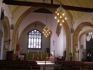 The inside of a church building. The foreground area shows a row of wooden benches. There is a large stained glass window in the background surrounded by plain white wall. Two suspended historic light fittings are shown, both lit up.