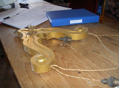 A gold painted church fitting laid out on a wooden table. The foreground shows the string that the fitting was suspended from. In the background there is a blue folder and some paperwork