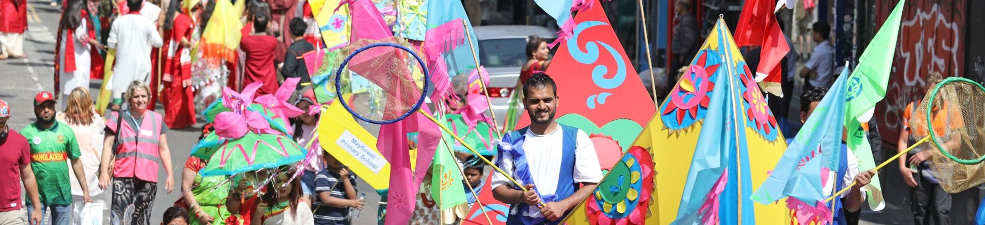 A carnival parade makes its way up a road. People wear colourful clothing, and carry flags. At the head of the procession a man walks inside a boat made of fabric stretched around a frame.