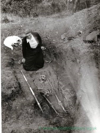 Dame Kathleen Kenyon pictured kneeling on the ground next to a skeleton during the excavation of the Sutton Walls. The photograph is taken from a height looking down on the grave.