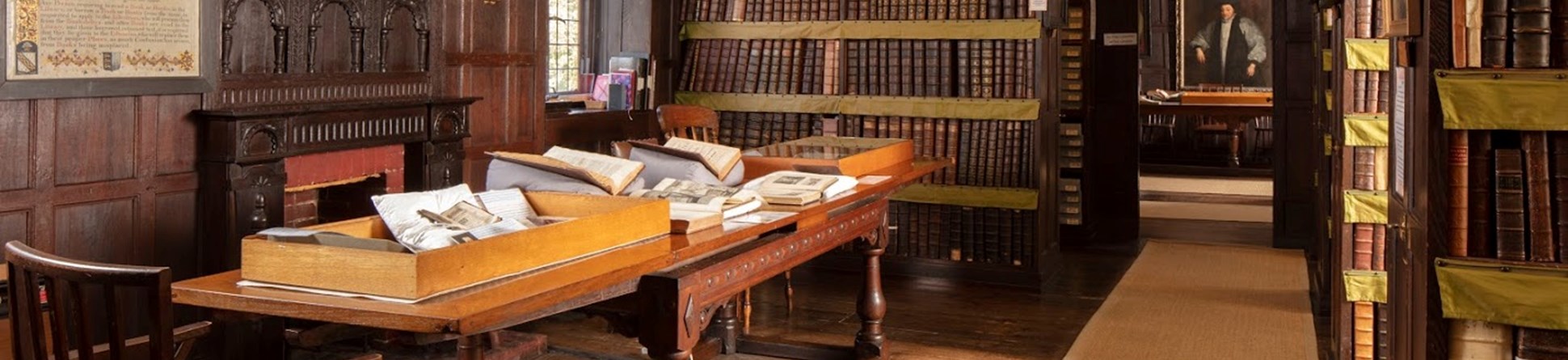 The library room shows dark wood panelling, wooden floorboard and dark wood bookcases with a long table to the left, covered in historic books. Historic portraits line the walls.