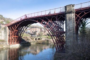 Red coloured iron bridge. There is a hill on the left side with old buildings, a grass bank and trees, and blue sky in the background. The river in the foreground is still with a reflection of the blue sky, the bank and the bridge. The bank on the right hand side features some shrubbery.
