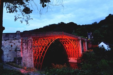 An iron bridge in a dusk setting. The bridge is lit up with red light and is surrounded by woodland and vegetation. There is a flowing river underneath the bridge, a traditional white building on the right and a cloudy dark blue sky in the background.