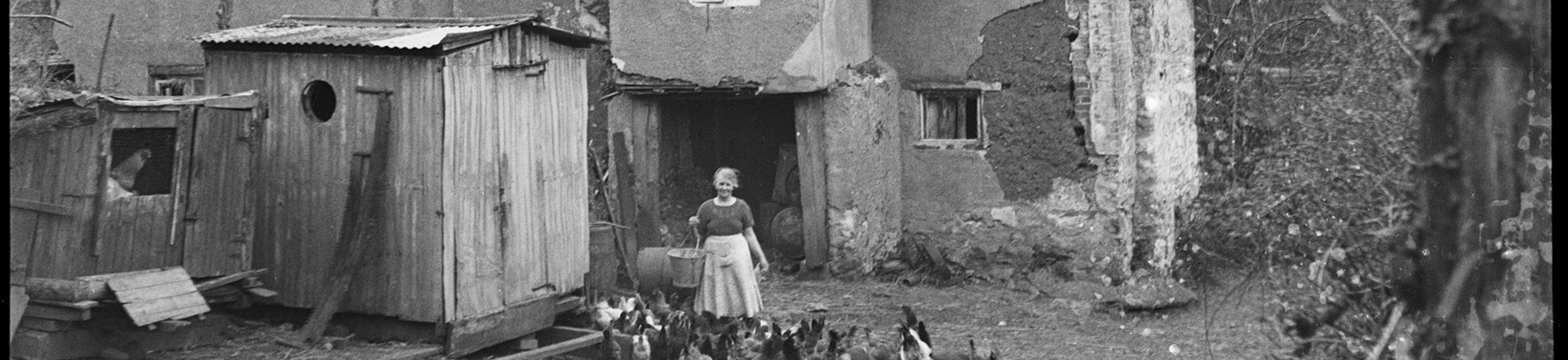 Black and white photograph of part of a rendered and thatched farmhouse. In the yard to the front, a woman holding a pail stands amongst a group of chickens. To her side is a shed or old shepherd's hut. Fields rise in the distance behind the farmhouse.