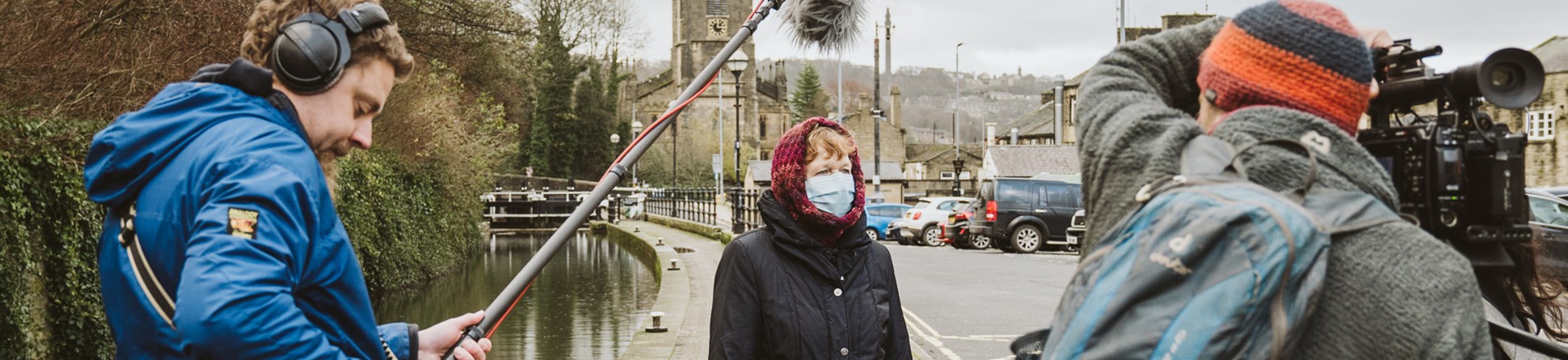 A woman wearing a mask is interviewed with a canal and church in the background. Camera crew are left and right in the foreground.