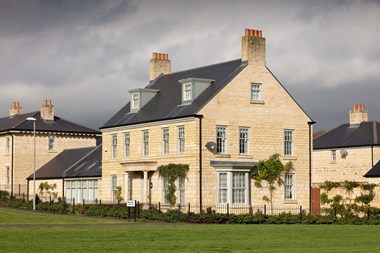 In the foreground is a modern detached house with two storeys plus attic level with dormers; stone exterior, symmetrical frontage with a central entrance portico to the ground floor; other dwellings in the background.