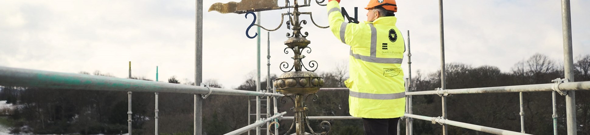 Heritage craftsman in a hard hat and high visibility jacket stands on scaffolding on the Wentworth Woodhouse, and reaches up to the weathervane. The sky behind him is cloudy.