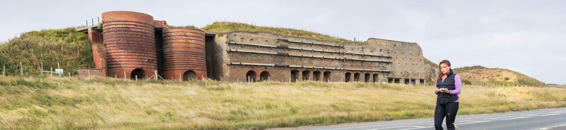 Stone- and brick-built battery structure with horizontal reinforcing timbers with a series of ground-level arched openings to the lime kilns it houses. Dune grass grows in front of it and grassy banks protrude above and either side of the structure.