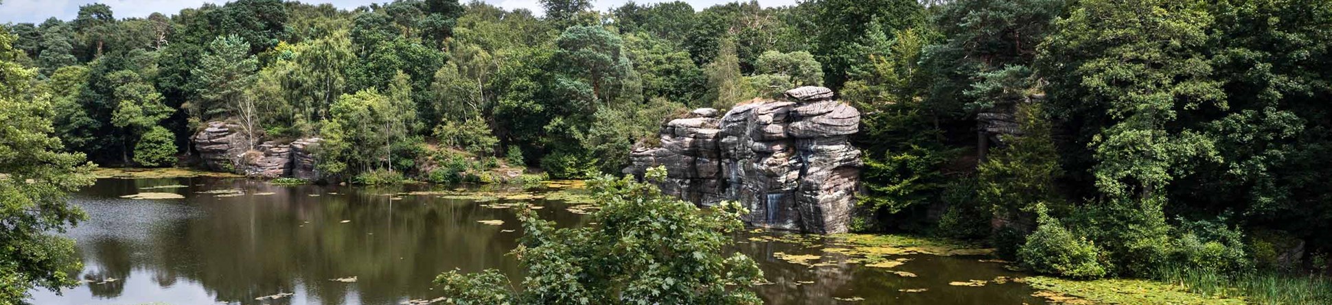 Landscape gardens, lake, rocks and dam wall at Plumpton Rocks. A man walks along the dam wall on the right of the image.