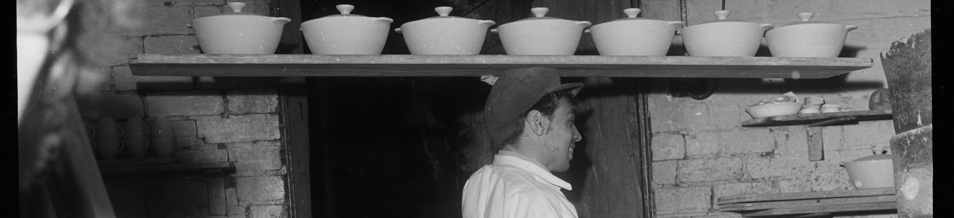 Man wearing cap and apron carries seven unglazed pots with lids on a plank on his head.