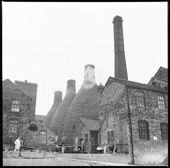Brick built pottery buildings, including three bottle kilns with five figures visible, going about their business just in front of the buildings.