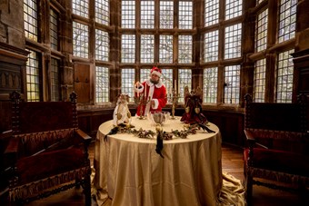 A person dressed in a Santa costume lights candles on a round table decorated with festive garlands, in front of some historic windows