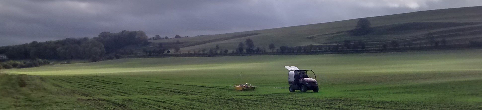 Colour photograph of buggy pulling a "sledge" across a field with germinating crop under a threatening sky heavy with clouds