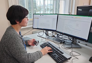 Photograph showing a short, dark-haired woman working at a desk, she is using two screens and is looking at a series of graphs while typing on the keyboard.