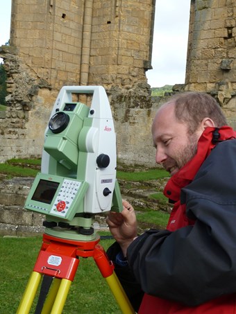 Colour photograph of a man using a total station theodolite with some ruined walls in the background