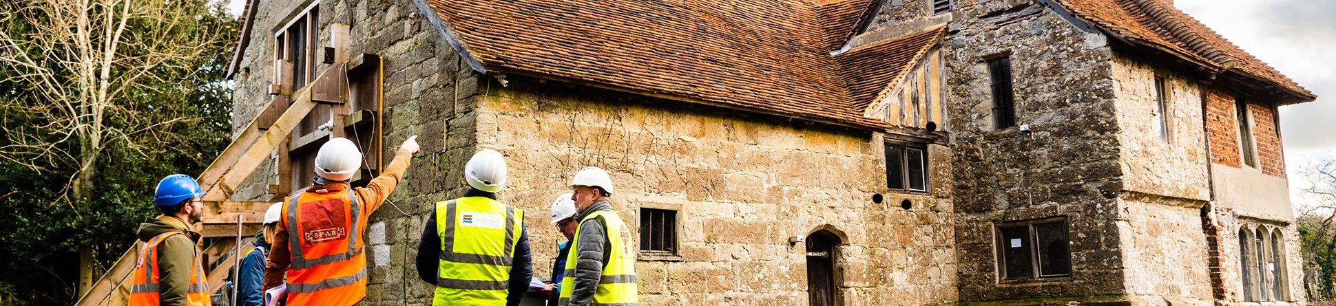A group of people in hard hats and high-vis jackets look at an old stone building.